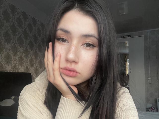 PattiTropea with long black hair, pouty pink lips, and freckles, hand on cheek, wearing a beige sweater, looking at camera, soft light, slightly flushed, sexy.