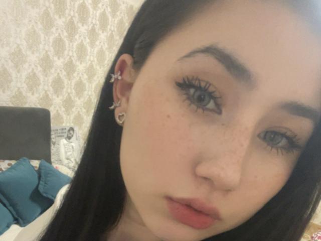 PattiTropea has pale skin, black hair, blue eyes, and wears a small ear ring with a butterfly. She has freckles, long eyelashes, and red lips, looking at the camera.