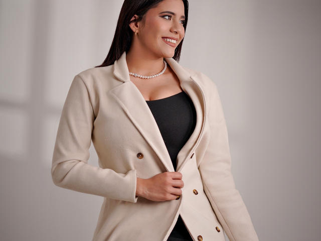 LucieMilner with long dark hair, smiling, wearing a beige double-breasted coat over a black top, pearl necklace, holding coat, tits visible under coat, soft light.