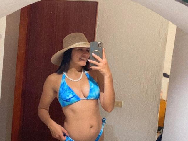 Lucie Milner, con un bikini azul, un sombrero y un collar de perlas, mostrando sus pechos y glúteos en un selfie frente a un espejo, sosteniendo un teléfono con una sonrisa encantadora..