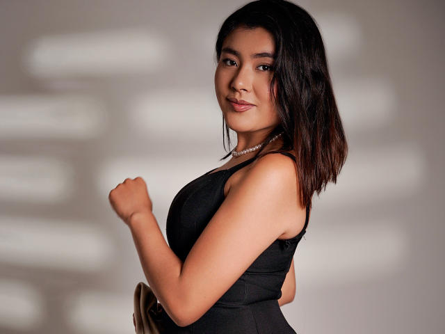 HannaGoez with medium tits in a black dress, brown hair, wearing a pearl necklace, smiling, raised fist, and brown purse, standing against a blurred white background.
