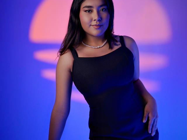 HannaGoez with dark hair, wearing a black sleeveless top and pearl necklace, standing with a slight smile, blue and pink light on her. Her tits are small and firm.