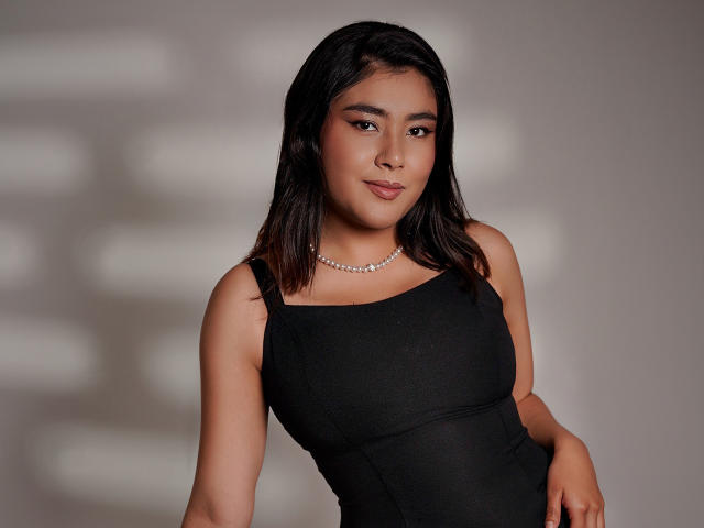 HannaGoez with medium brown skin, black wavy hair, wearing a black dress, pearl necklace, and subtle makeup, has a confident, slightly smug expression, and is showing her tits.