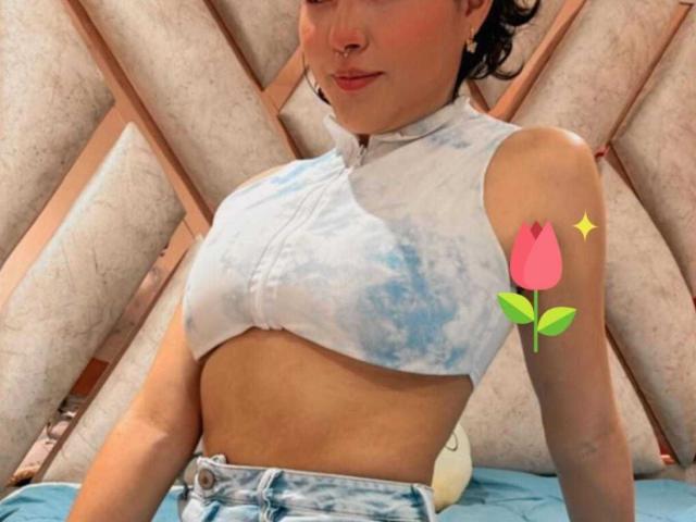 HannaGoez with small nose ring, wearing a tie-front, blue-white tie-dye crop top, showing her tits, and blue jeans, has a pink rose sticker on her right arm.