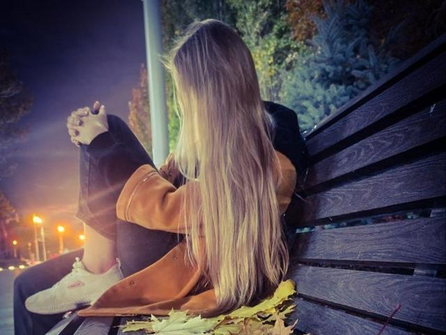 Tomiko Helfritz sitzt auf einer hölzernen Bank mit langen blonden Haaren, trägt einen braunen Mantel und schwarze Jeans, die Beine sind angezogen, die Hände auf den Knien, im Herbstlaub, Nacht, Straßenlaternen..