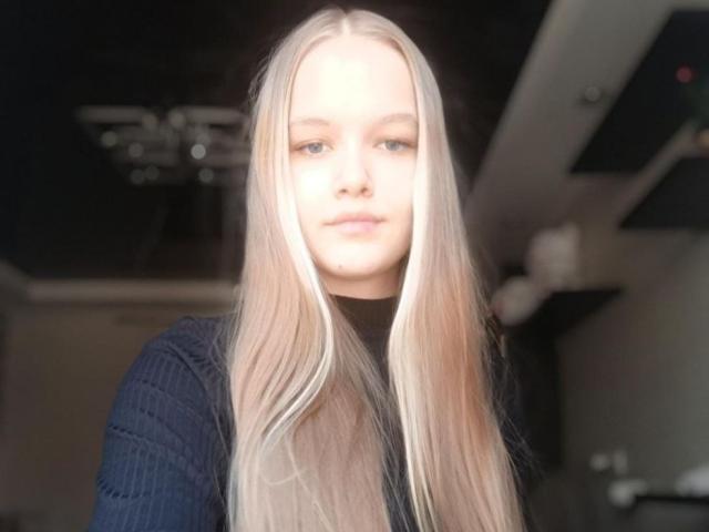 Tomiko Helfritz hat langes, glattes blondes Haar, helle Haut und ein leichtes Lächeln. Sie trägt ein schwarzes, geripptes Oberteil mit langen Ärmeln. Ihre blauen Augen sind leicht verschwommen..
