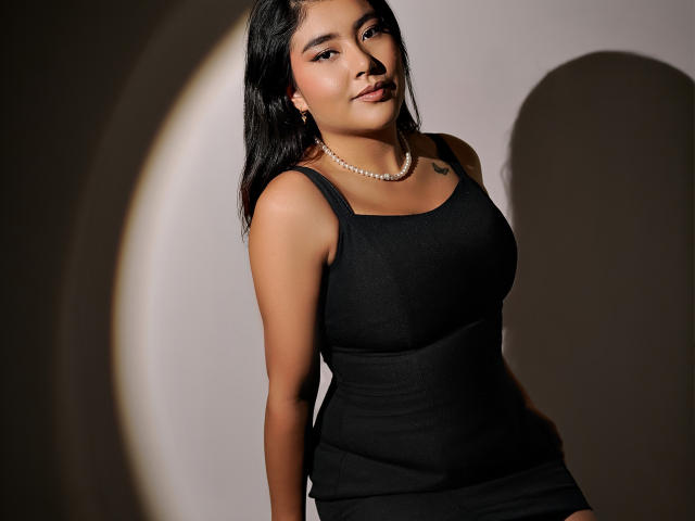 HannaGoez with tan skin, black tank top, and pearl necklace, showing her tits, dark hair, and a slight smile, standing against a shadowed background.