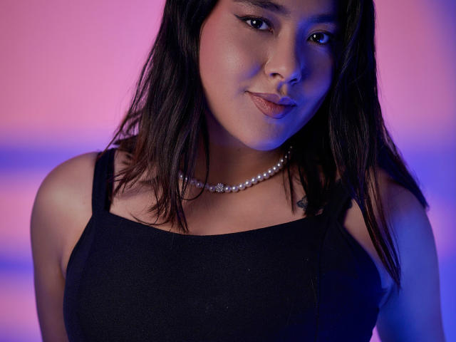 HannaGoez with dark brown hair, wearing a black tank top, pearl necklace, and subtle makeup, looking slightly to the side with a gentle smile, in soft blue and pink light.