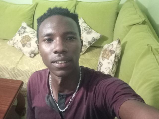 Ebonyd, com pele escura, cabelo curto e encaracolado, vestindo uma camisa marrom e uma corrente de prata, sentada em um sofá verde com almofadas florais, sorrindo para a câmera..
