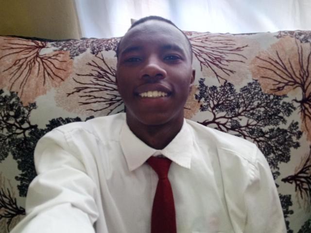 Ebonyd tem a pele escura, cabelo curto, veste uma camisa branca com uma gravata vermelha, está sorrindo mostrando os dentes e sentada em um sofá com estampa floral..