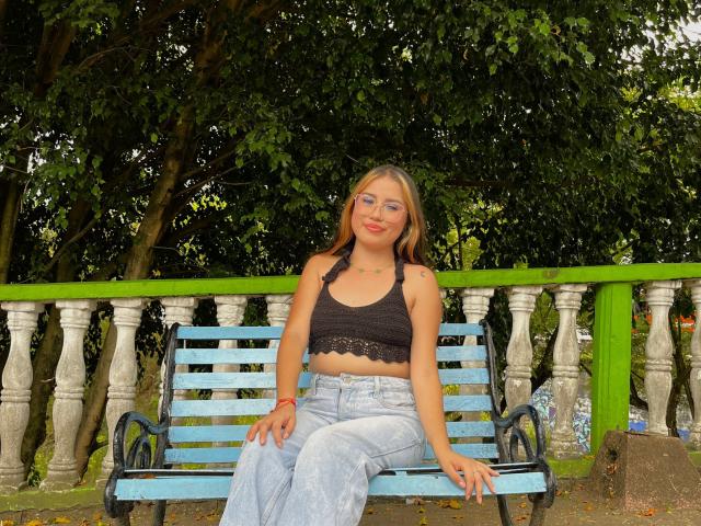 AmilyRizzo is sitting on a blue bench, wearing a black lace crop top and light blue jeans, with glasses and red lipstick, showing her small tits and a tattoo.