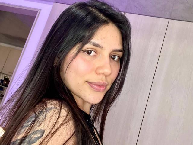 SophiaMarx with long black hair, brown eyes, light pink lips, subtle tattoos on her right shoulder, smiling at the camera, wearing a black top, in a brightly lit room.