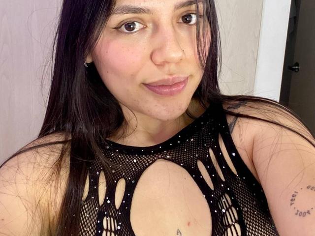 SophiaMarx, con lunghi capelli castani, indossa una maglia nera in rete con ampi spacchi che mostrano il seno, sorride, ha le labbra rosa, un piccolo tatuaggio sul braccio destro, e sullo sfondo si vede una porta bianca..