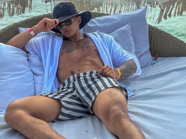 PaulALLEN, sin camisa, con pantalones cortos a rayas negras y blancas, una camisa azul abierta, un sombrero negro, gafas de sol y brazaletes, recostado en una cama de mimbre con almohadas grises..