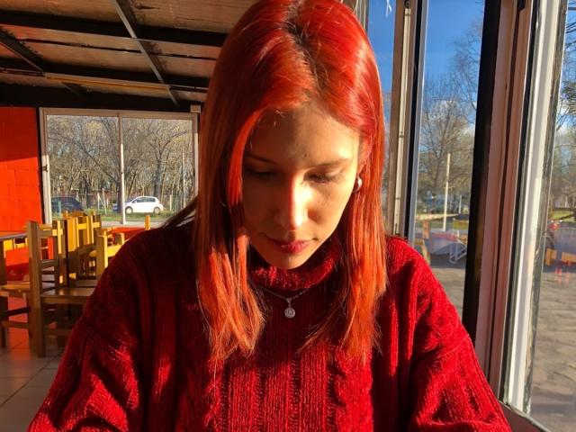 KiaraHernandezX with red hair, red knit sweater, looking down, sunlight on her face, sitting by window, visible reflection, wooden chairs in background, calm expression, soft light.