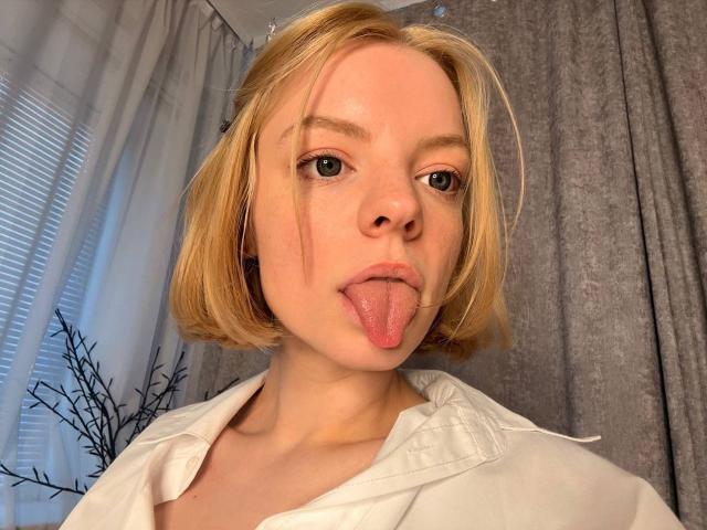 MyrleMogush has blonde bob, sticking out tongue, wearing white shirt, small hoop earrings, fair skin, light brown eyes, and a slightly flushed face in a selfie.