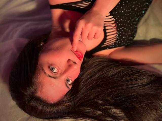 SaraGraham's face is upside down with long black hair, wearing a black, cut-out dress, her finger touching her red-lipped, slightly parted lips, looking up with green eyes.