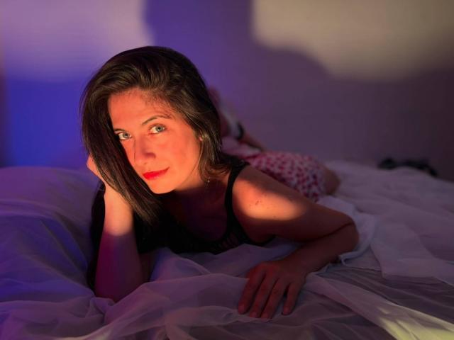 SaraGraham with red lips, black tank top, lying on white bed, arms resting, shadowed by blue and orange light, smiling, brown hair, hand on cheek, soft lighting.