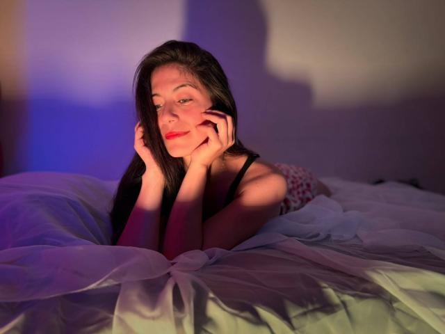 Sara Graham, com longos cabelos pretos, batom vermelho e as mãos nas bochechas, deitada em uma cama com lençóis brancos, suavemente iluminada por sombras roxas e azuis..