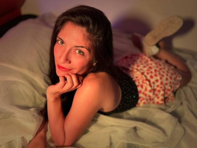 SaraGraham with green eyes, lying on a bed, wearing a black top and red patterned skirt, hands under chin, smiling, brown hair, white sheets, white shoes.