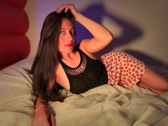 SaraGraham with long brown hair, wearing a black sleeveless top and red heart-patterned shorts, lying on a bed with a hand in her hair, looking at the camera.