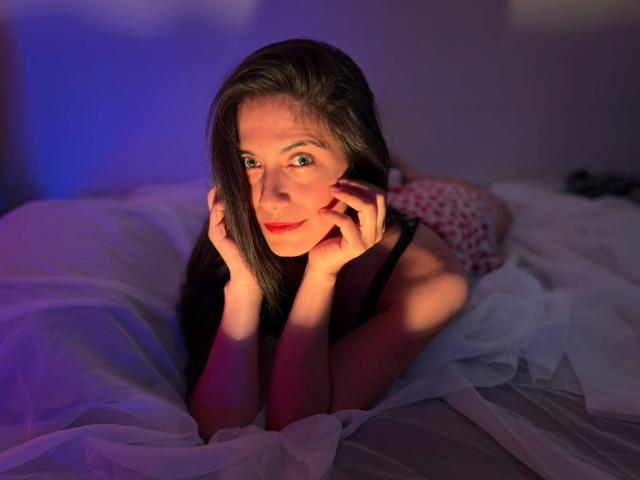 Sara Graham, deitada em uma cama, com as mãos nas bochechas, tem cabelos castanhos longos, olhos verdes, lábios vermelhos e está vestindo uma blusa preta, sob iluminação roxa e azul..