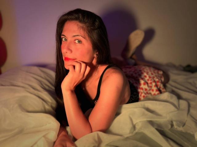 Sara Graham, com cabelos castanhos longos, batom vermelho, blusa preta de renda, deitada na cama, apoiada no cotovelo, mão no queixo, lençóis brancos, shorts com estampa vermelha e branca, iluminação de quarto..