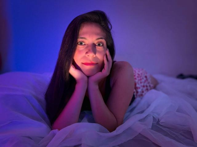 Sara Graham, com cabelos longos e escuros, mãos nas bochechas, deitada em uma cama branca, com iluminação azul e roxa, sorriso suave, olhando para a câmera, vestindo um vestido floral vermelho, relaxada, fofa..