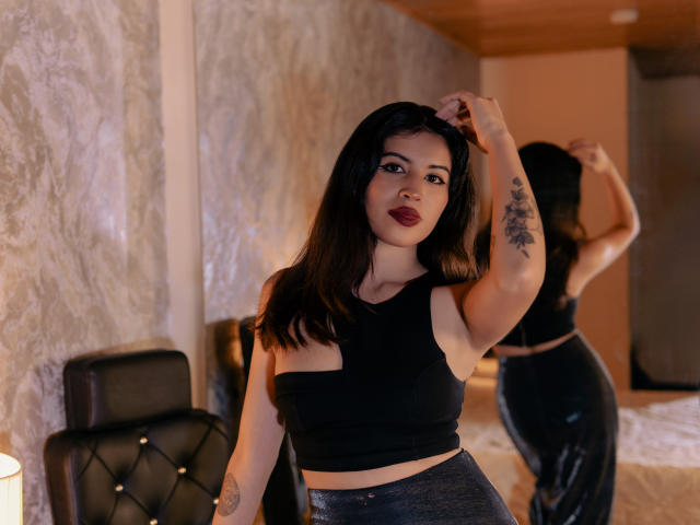 TifanyLopez with dark hair, red lipstick, black crop top, and shiny pants, standing in front of a mirror, hand in hair, tattoo on arm, reflection showing her ass.
