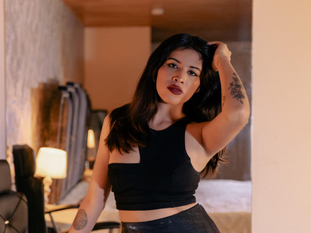TifanyLopez with dark hair, wearing a black crop top, exposing her small tits, has a tattoo on her arm, hand in hair, standing in a warmly lit room.