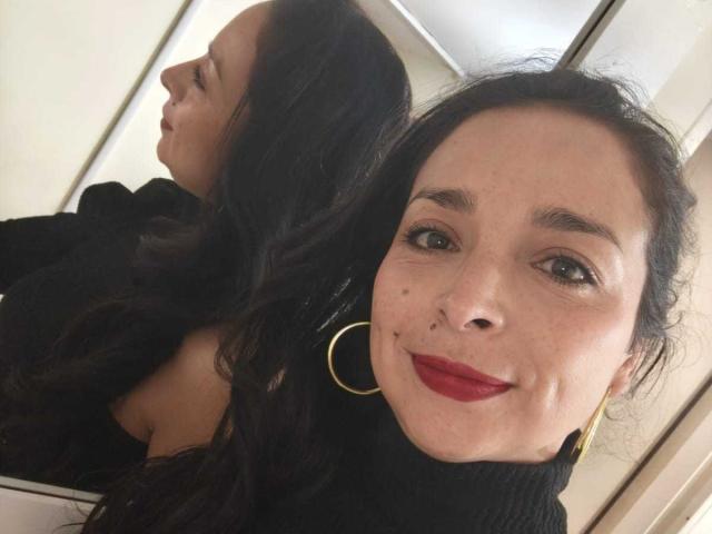 StangelarRivera, con lunghi capelli neri, rossetto rosso, un maglione nero a collo alto e orecchini a cerchio dorati, sorride allo specchio, mostrando la sua schiena e il sedere scoperto. Specchio riflettente a sinistra..