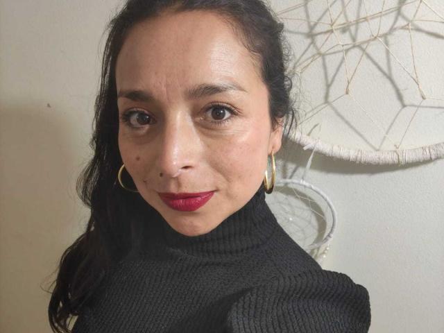 StangelarRivera, con capelli scuri, rossetto rosso, un maglione nero a collo alto, orecchini a cerchio dorati e una piccola lentiggine sul lato destro del viso. Le sue natiche non sono visibili..
