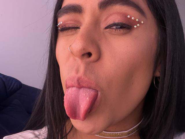 StellaBeckker's face with dark hair, brown eyes, and nose ring. She's winking with pink glittery eyeliner, gold choker, and sticking out her pink tongue.