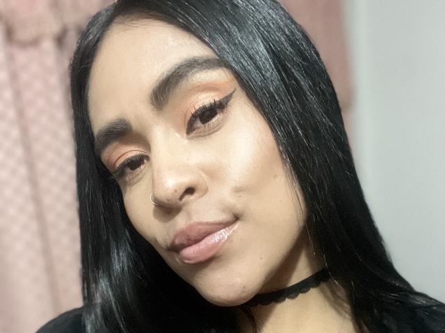 StellaBeckker with shiny black hair, nose ring, and choker, wearing orange eyeshadow, winged eyeliner, and pinkish glossy lips, looking down with a subtle smile.