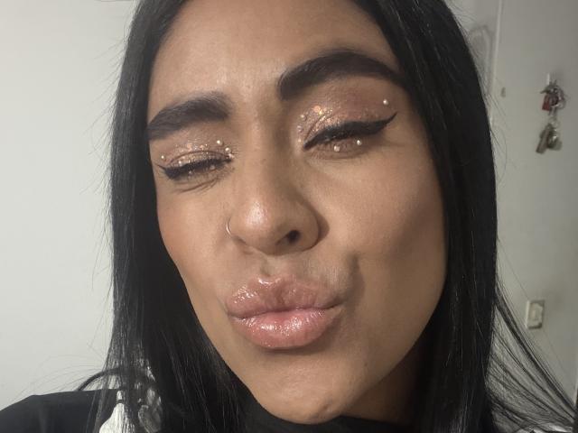 StellaBeckker's face with glossy pink lips, black eyebrows, brown glittery eyeshadow, and a nose ring, pouting slightly, with long black hair, in a selfie.