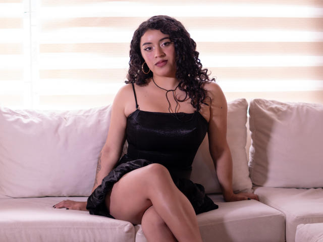 AlinnaPalacios with curly black hair, wearing a black spaghetti-strap dress, sitting on a white couch, legs crossed, looking at the camera, showing her ass, small gold hoop earrings.