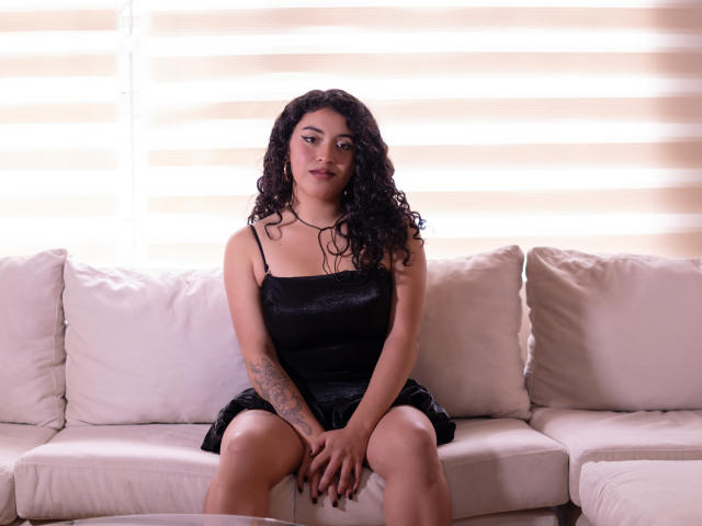 Alinna Palacios mit lockigem, schwarzem Haar, trägt ein schwarzes Kleid, sitzt auf einem weißen Sofa, die Hände auf den Knien, hat ein Tattoo am rechten Arm und blickt mit einem leichten Lächeln in die Kamera..