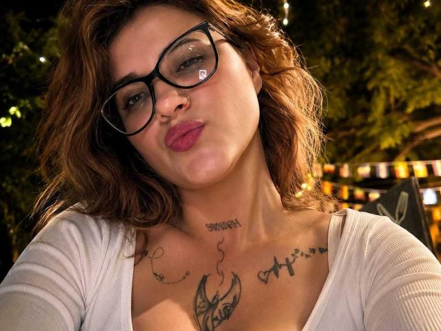 Antonella Tomsson, con gafas, labios rosados y fruncidos, cabello castaño ondulado, blusa blanca, tatuajes en el pecho y el cuello, pezones prominentes, iluminación tenue, fondo con árboles..