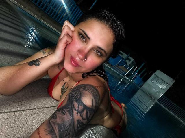 AntonellaTomsson, wet, red bikini, lying by the pool at night, hand on cheek, tattoos on arm and shoulder, dark hair, nose ring, smiling, water reflecting on concrete.