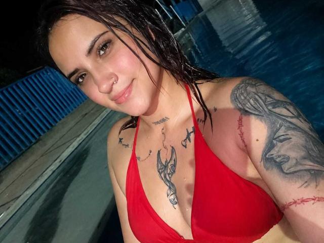 Antonella Tomsson, con i capelli castani bagnati, un top da bikini rosso e tatuaggi, inclusi un grande tatuaggio nero sul braccio destro e tatuaggi sul petto, sorride in una piscina di notte..