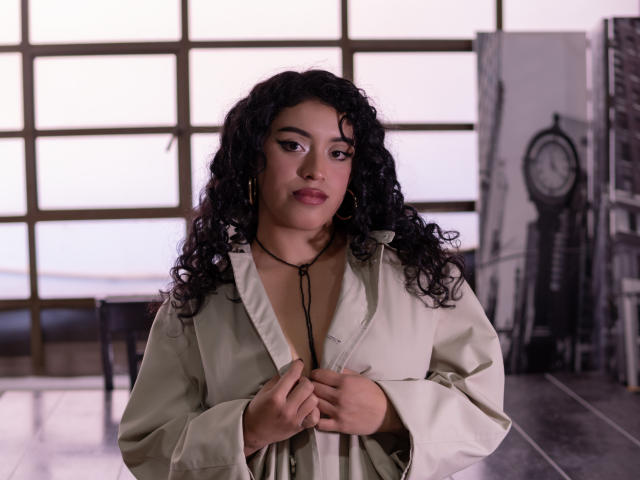 AlinnaPalacios with curly black hair, light brown skin, wearing a beige jacket, hands on chest, small black necklace, gold earrings, looking down with a subtle smile, indoor setting.