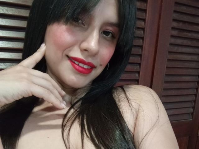 MadissonFontaine has black hair, red lipstick, pink blush, and is smiling with her finger on her cheek. She's topless with her tits partially visible. Her ass is not shown.