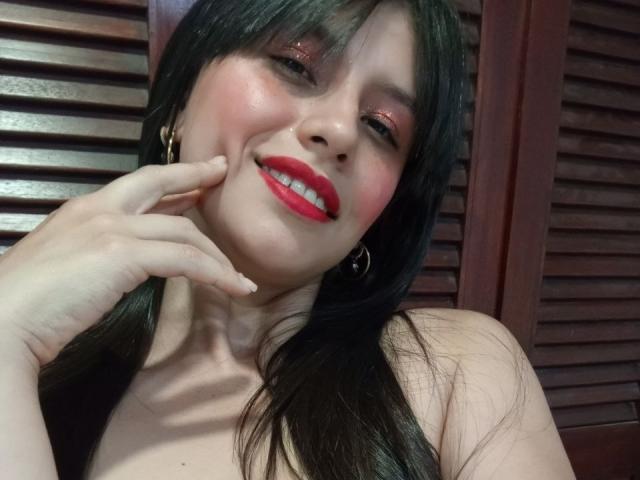 MadissonFontaine with long black hair, red lipstick, and pink eyeshadow, smiling with her hand near her face, showing her teeth. She's wearing gold hoop earrings, and she's topless.