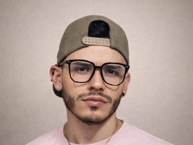 Lianferrer with a brown baseball cap, black glasses, beard, and stud earrings, wearing a pink shirt and gold necklace, looking serious.