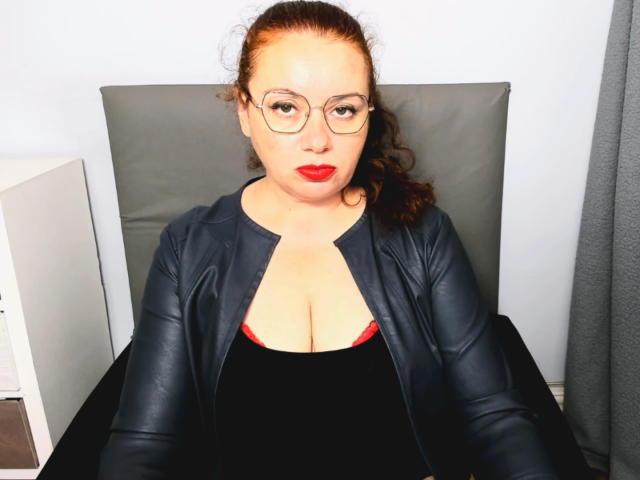 ElisabethLovee, com cabelo castanho encaracolado, batom vermelho, óculos, jaqueta de couro preta e blusa preta, mostrando seus seios grandes, sentada em uma cadeira, olhando para a câmera..