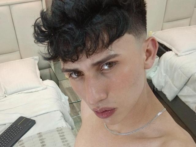 ThomaMees heeft kort, krullend zwart haar, bruine ogen en een zilveren ketting. Hij kijkt neutraal in de camera, is naakt en ligt in een bed met witte kussens..