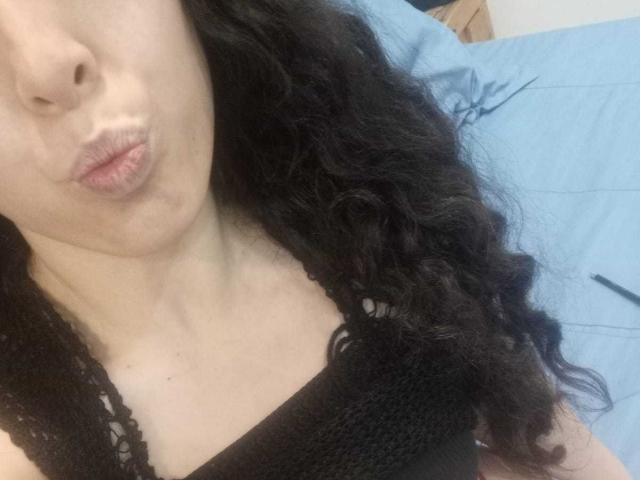 KendraSimons with puffy lips, black curly hair, wearing a black lace tank top, showing a hint of her tits, on a blue bed. Her nose is pierced.