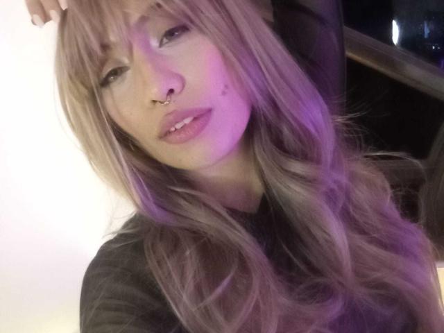 Ilaynna, con cabello rubio largo y ondulado, un piercing en la nariz y lápiz labial rosa, labios ligeramente entreabiertos y un brillo púrpura en su rostro, vestida con una camisa oscura, mirando a la cámara..