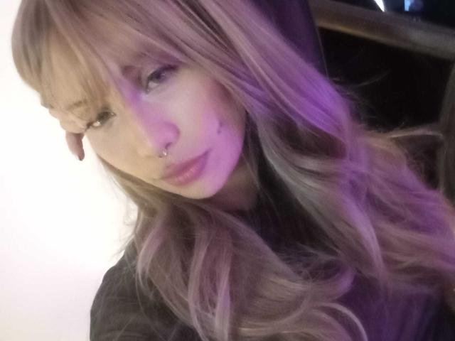 Ilaynna, con lunghi capelli ondulati di colore rosa-marrone, un anello al naso e rossetto rosa, tiene la testa con una luce viola tenue sul lato sinistro. Espressione dolce e sensuale..