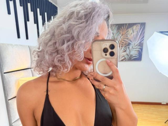 Elena Saenz, com cabelos brancos encaracolados, usando um top preto de alças, mostrando seus seios em uma selfie no espelho, segurando um anel de ouro e um celular com um suporte para "ring light"..